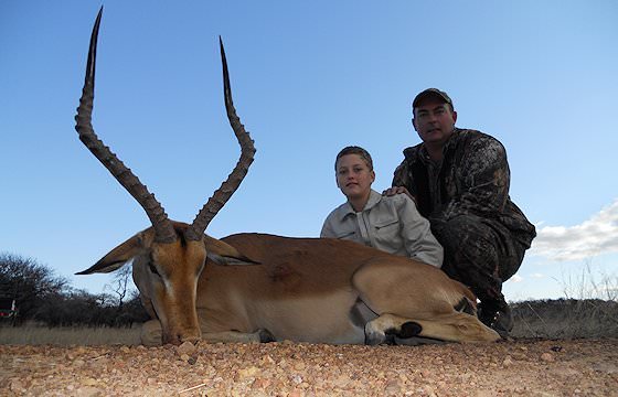 A professional hunter sits alongside a young hunter and his impala trophy.